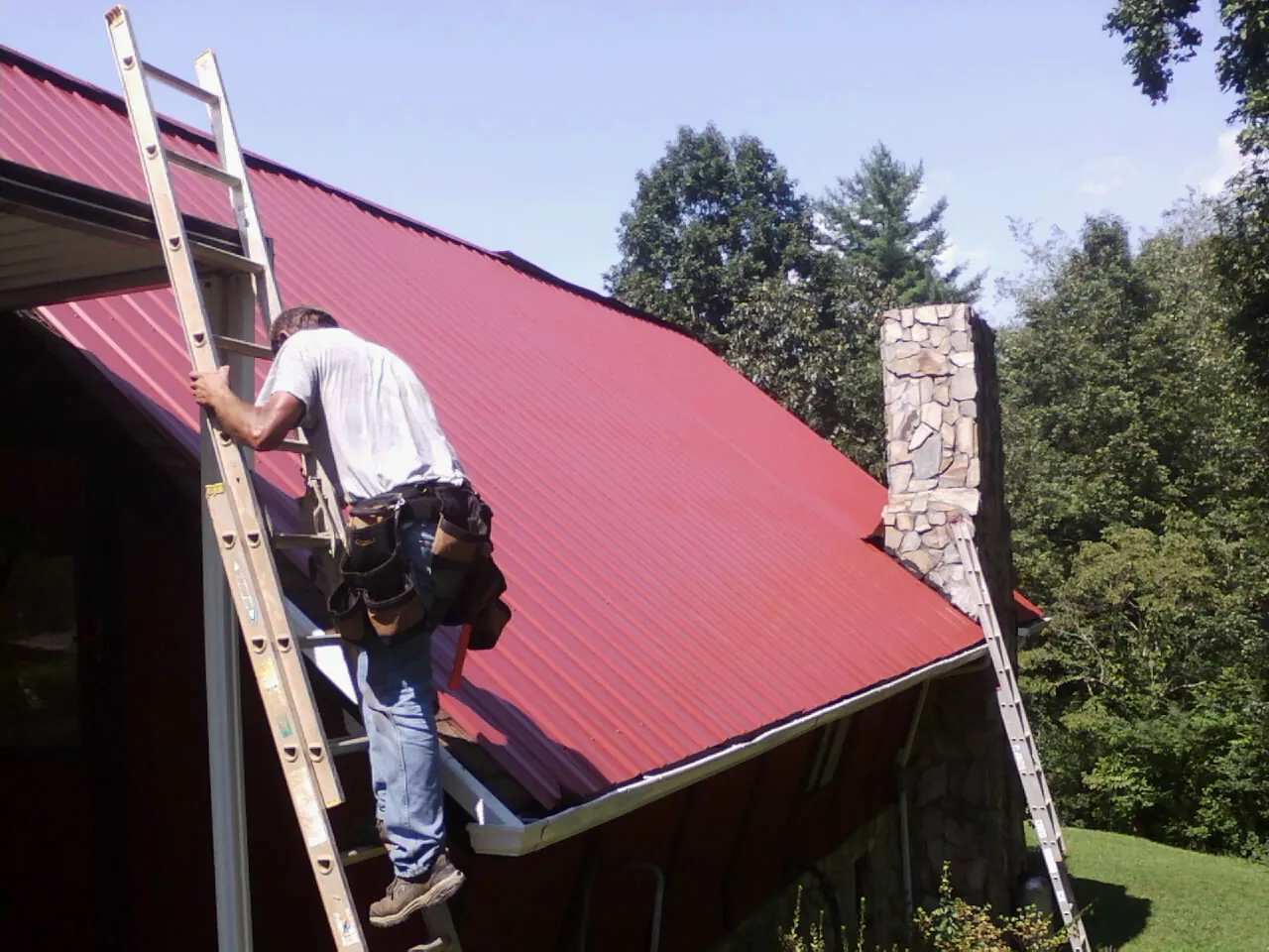 Roofer on a ladder beside a red metal roof and stone chimney — Soffit & Fascia Repair by Roofing Brothers in Ethel